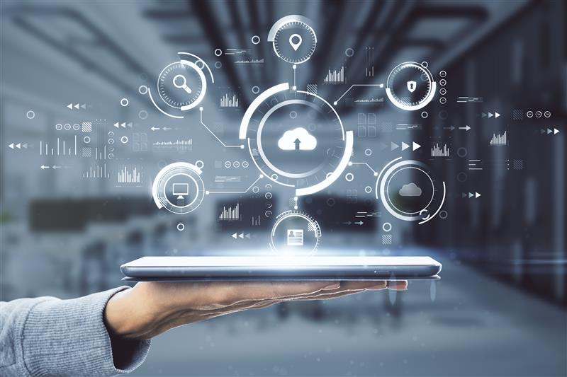 A hand holds a tablet with digital cloud icons and data graphics floating above in a modern, blurred office setting. Blue-white color scheme.