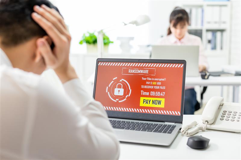 Man frustrated at desk with a laptop showing ransomware warning in red. "Pay Now" button visible. Woman working in background. Office setting.