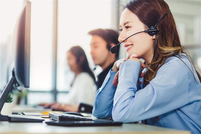 Woman smiling at computer, wearing headset in bright office, with two colleagues in background. Professional and cheerful atmosphere.