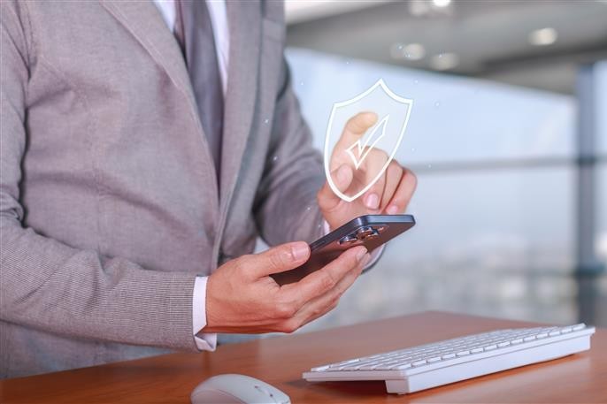 Businessperson in a suit using a smartphone with a digital shield icon. Office setting with a keyboard and mouse on a wooden desk.