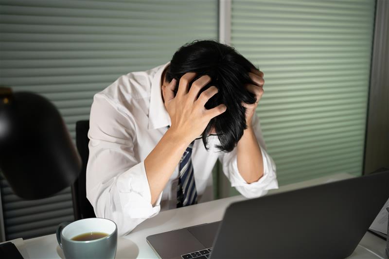 Person in white shirt and tie sits at a desk, head in hands, appearing stressed. A laptop, lamp, and coffee cup are on the desk.