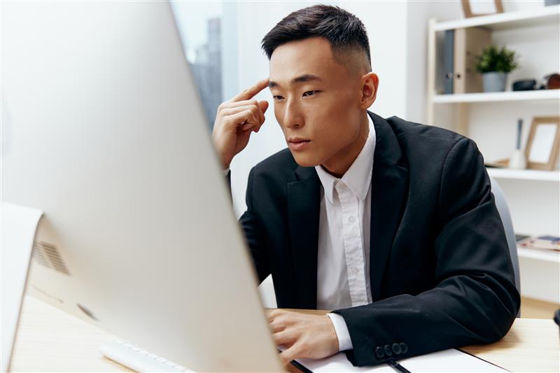 Man in a black suit focuses on a computer screen in a modern office, touching his temple thoughtfully. Bright, organized shelves in the background.