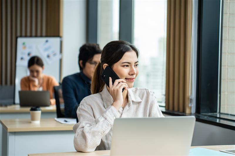 A woman smiling on the phone in an office, sitting at a desk with a laptop. Others work in the background. Bright, modern setting.