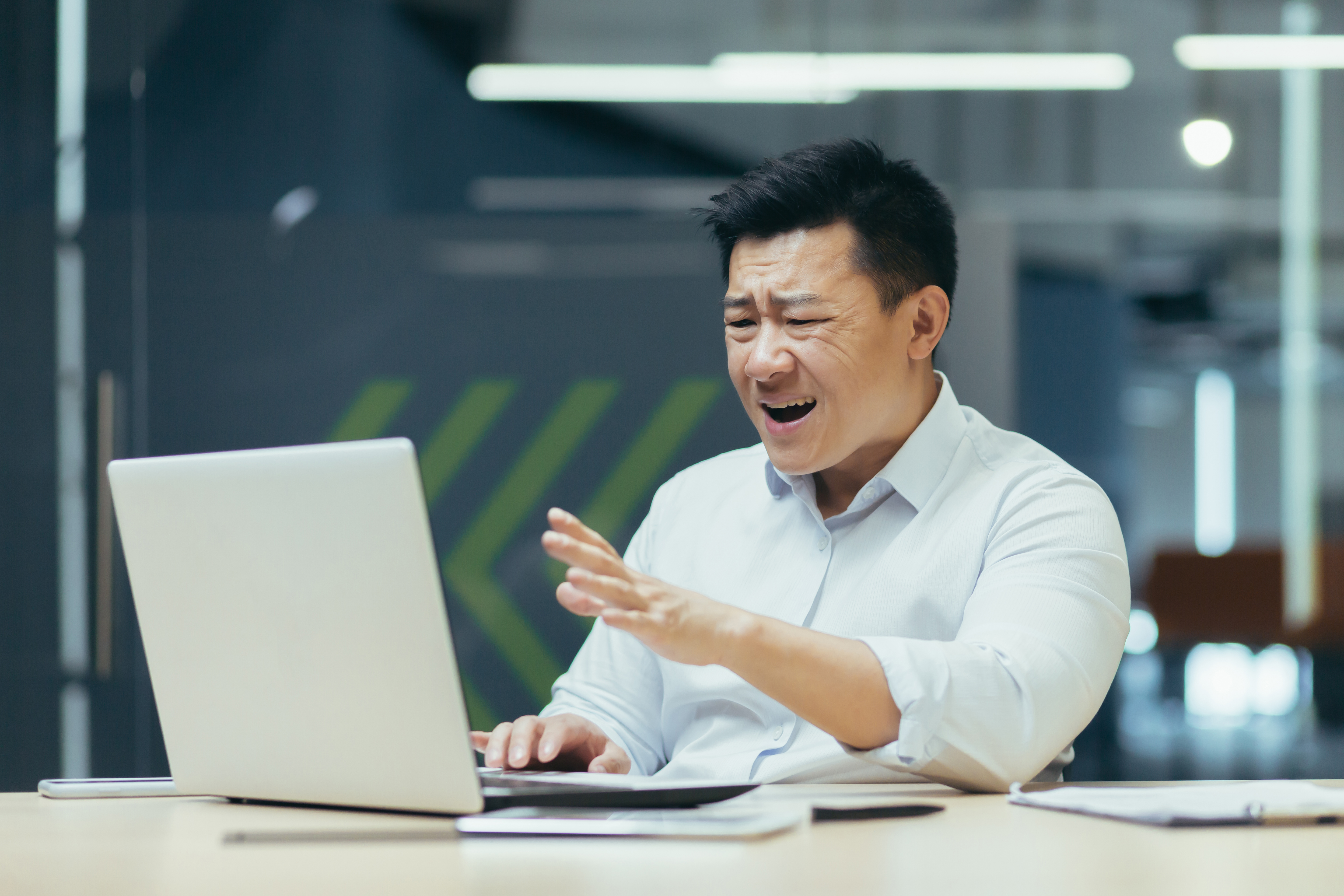 Man in white shirt at desk reacts intensely to a laptop screen. Office setting with blurred background and diagonal green stripes.
