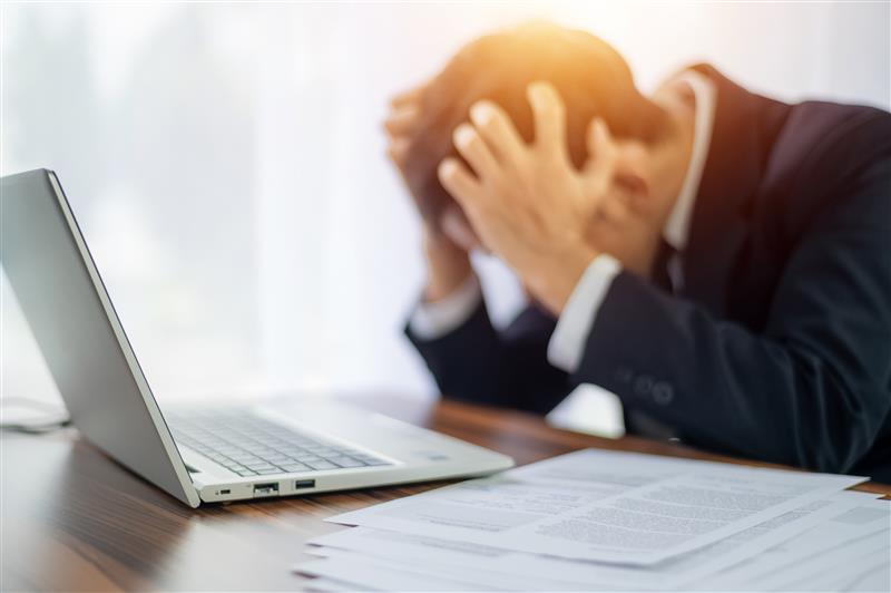 Businessperson in suit, head in hands, sits at desk with laptop and scattered papers, conveying stress. Bright light from window.