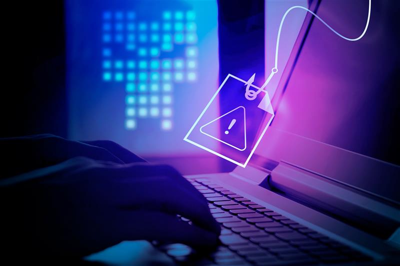 Hands typing on a laptop, a phishing symbol displayed. Background shows a digital threat icon in blue and purple lighting.