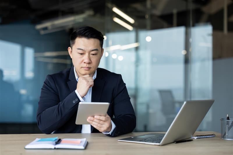 Man in a suit sits at a desk in a modern office, reading a tablet. A laptop, notebook, and pen are nearby. He appears thoughtful.