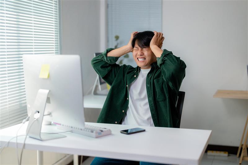 Person in a green shirt at a desk with a computer, holding their head in frustration. Bright room with blinds and a smartphone on the desk.