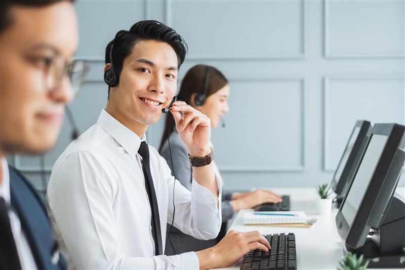 Smiling call center employees at desks with headsets, working on computers in a modern, light gray office setting.