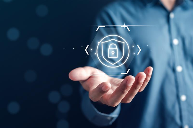 A person holds a glowing digital lock and shield icon in their palm, symbolizing security. The background is dark blue with soft light spots.