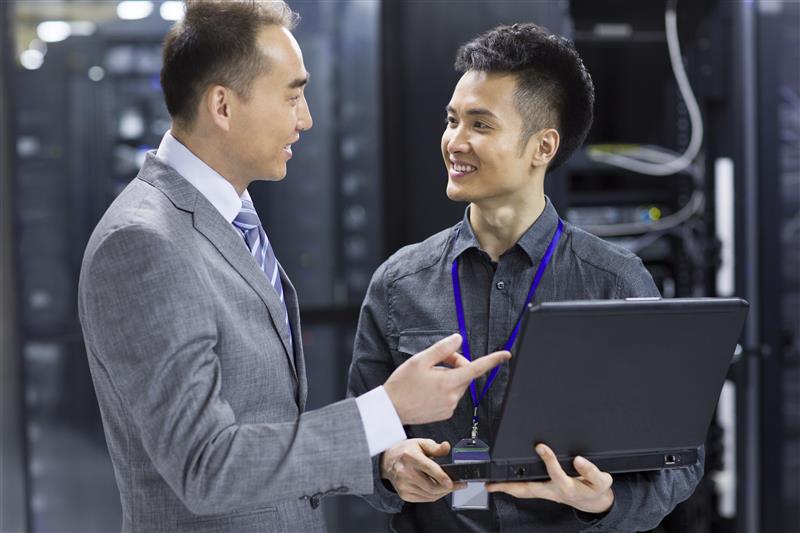 Two men in a server room, one in a suit pointing, the other holding a laptop. Both smiling, suggesting a successful discussion.
