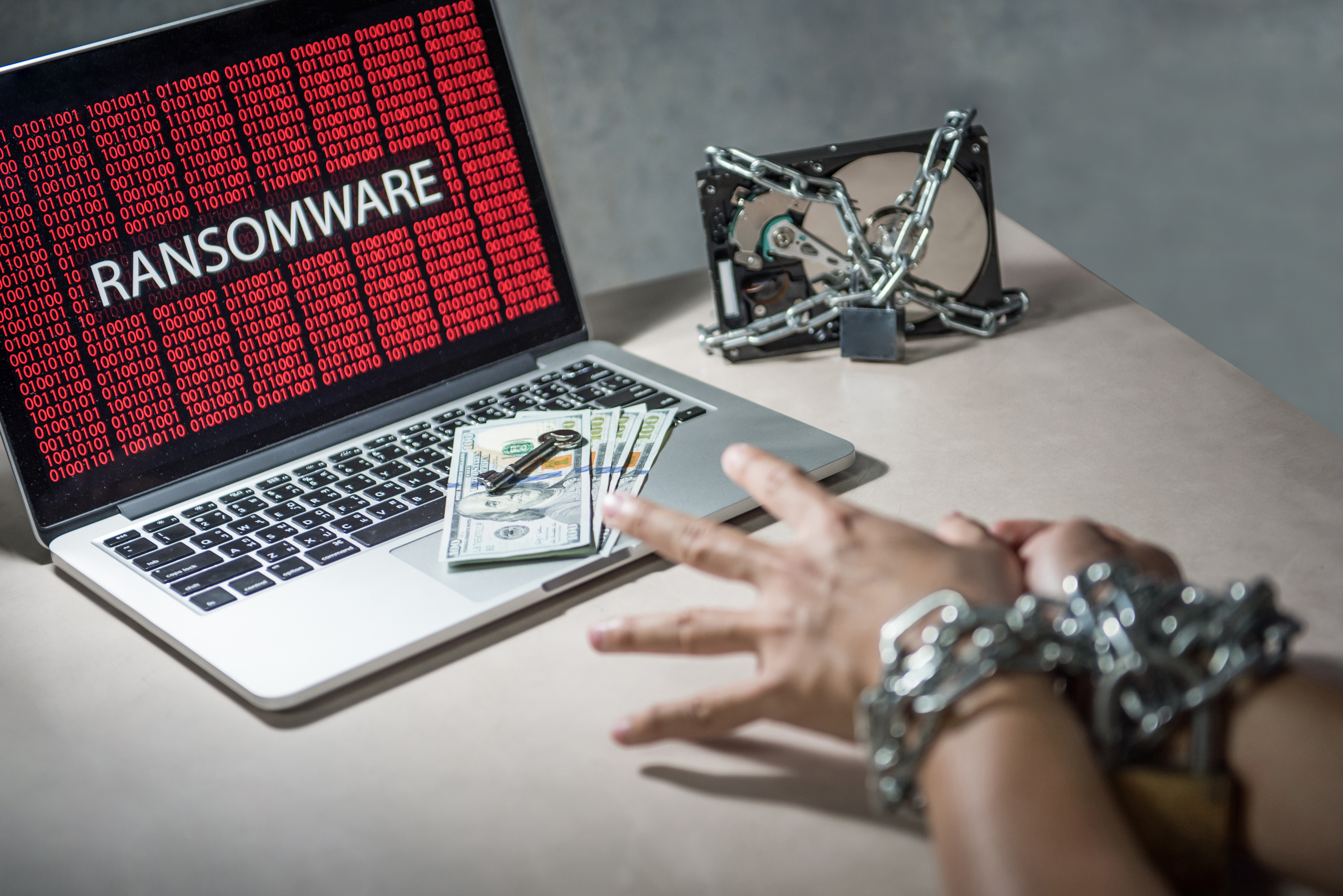 Laptop showing "Ransomware" in red, handcuffed hands reaching for money and key on the keyboard, chained hard drive in the background.