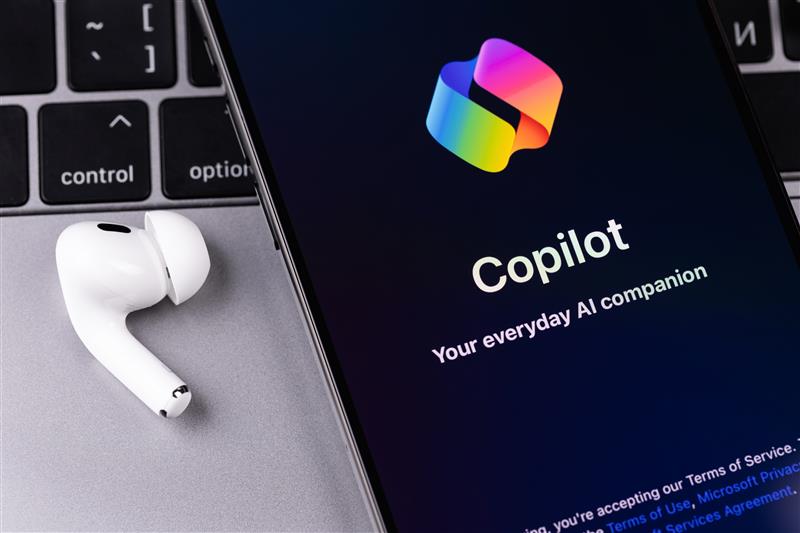 Smartphone displaying "Copilot, Your everyday AI companion" beside a white earbud on a silver laptop keyboard.