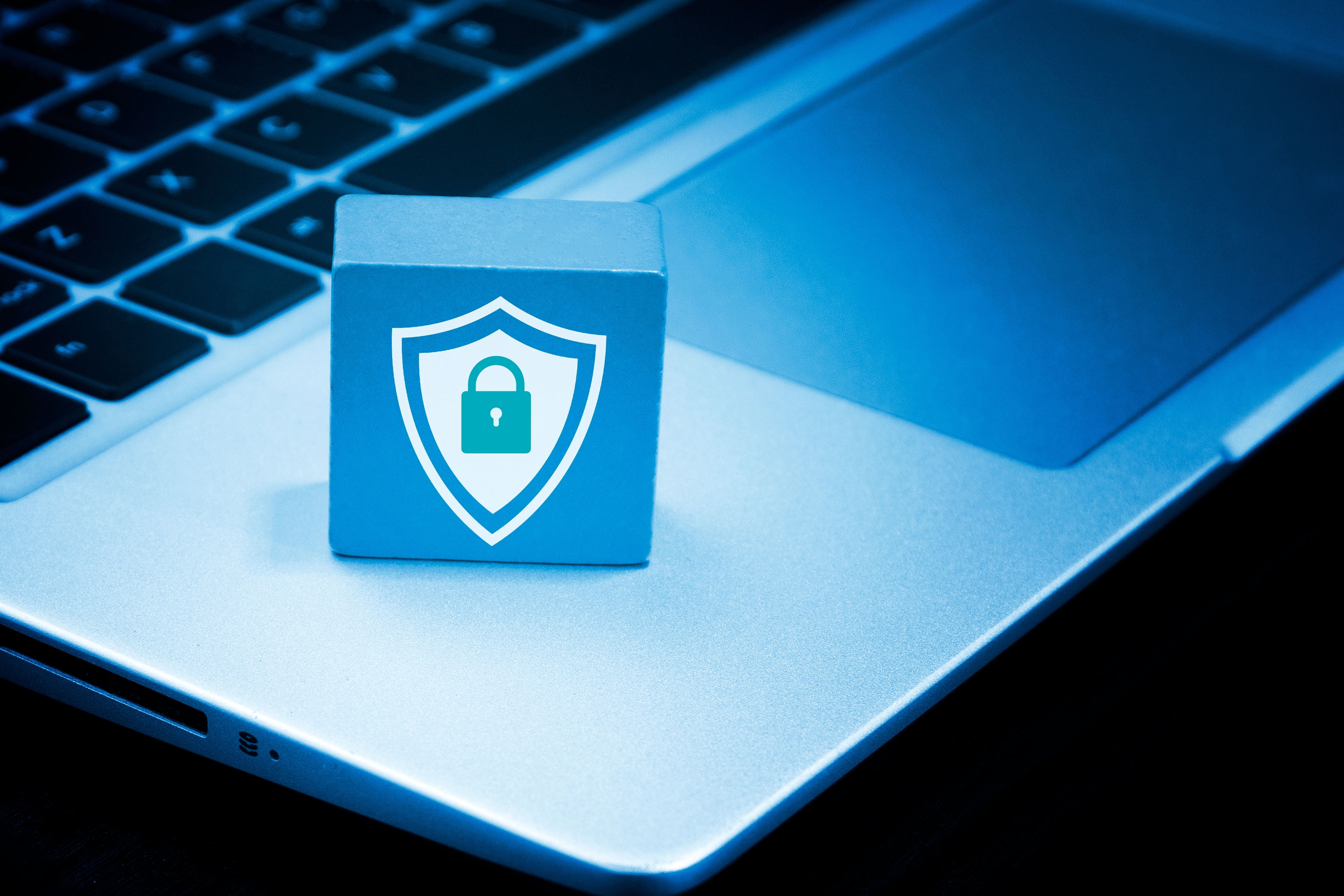 Blue padlock icon on a cube, placed on a gray laptop. The background has a soft blue tone, symbolizing data security.
