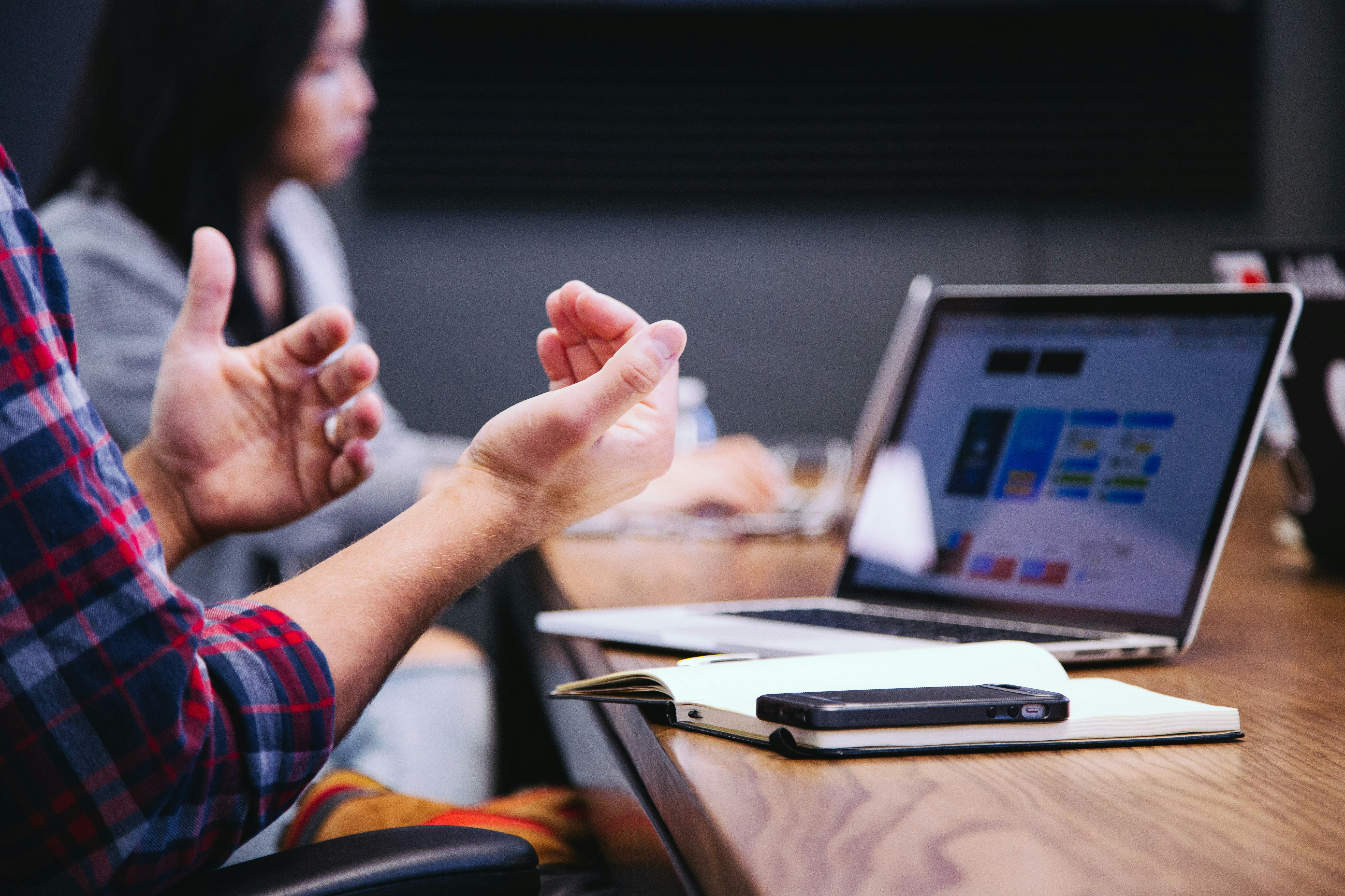 Person gesturing at a meeting table. Open laptop with charts visible. Notebook and phone on table. Focused, collaborative setting.