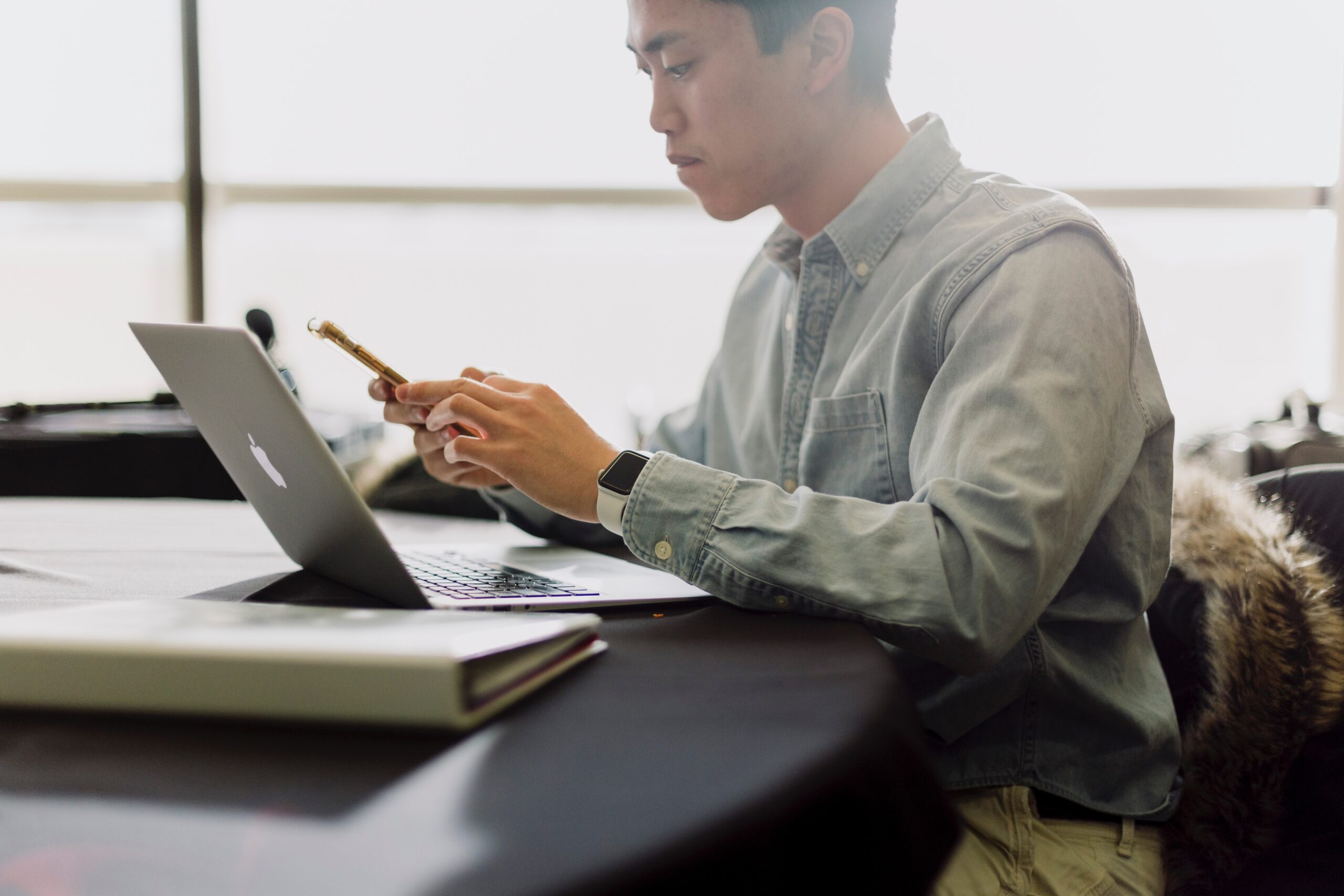 A person in a denim shirt looks at a phone while sitting at a table with a laptop. Soft, natural light fills the room. Calm mood.